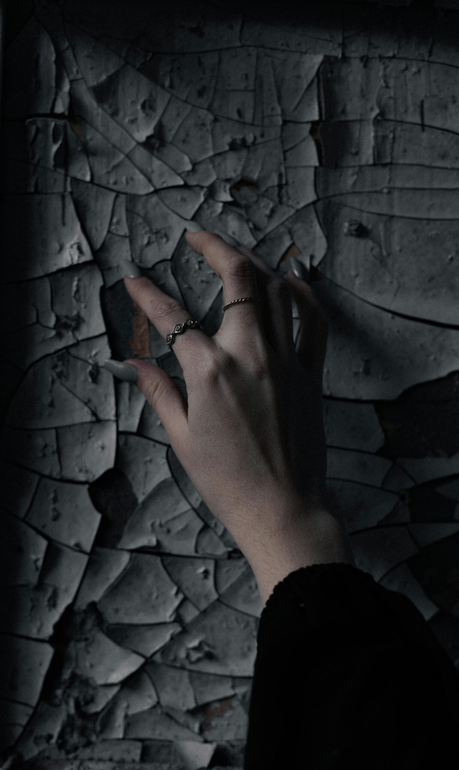 A hand with manicured nails and rings gracefully touching a cracked wall, creating a moody and artistic scene.