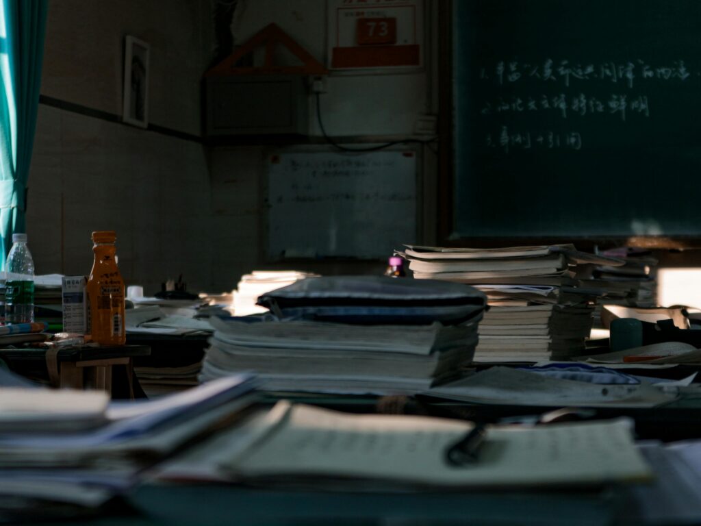 Messy tables with big heaps of papers and stationery in dark room with blackboard