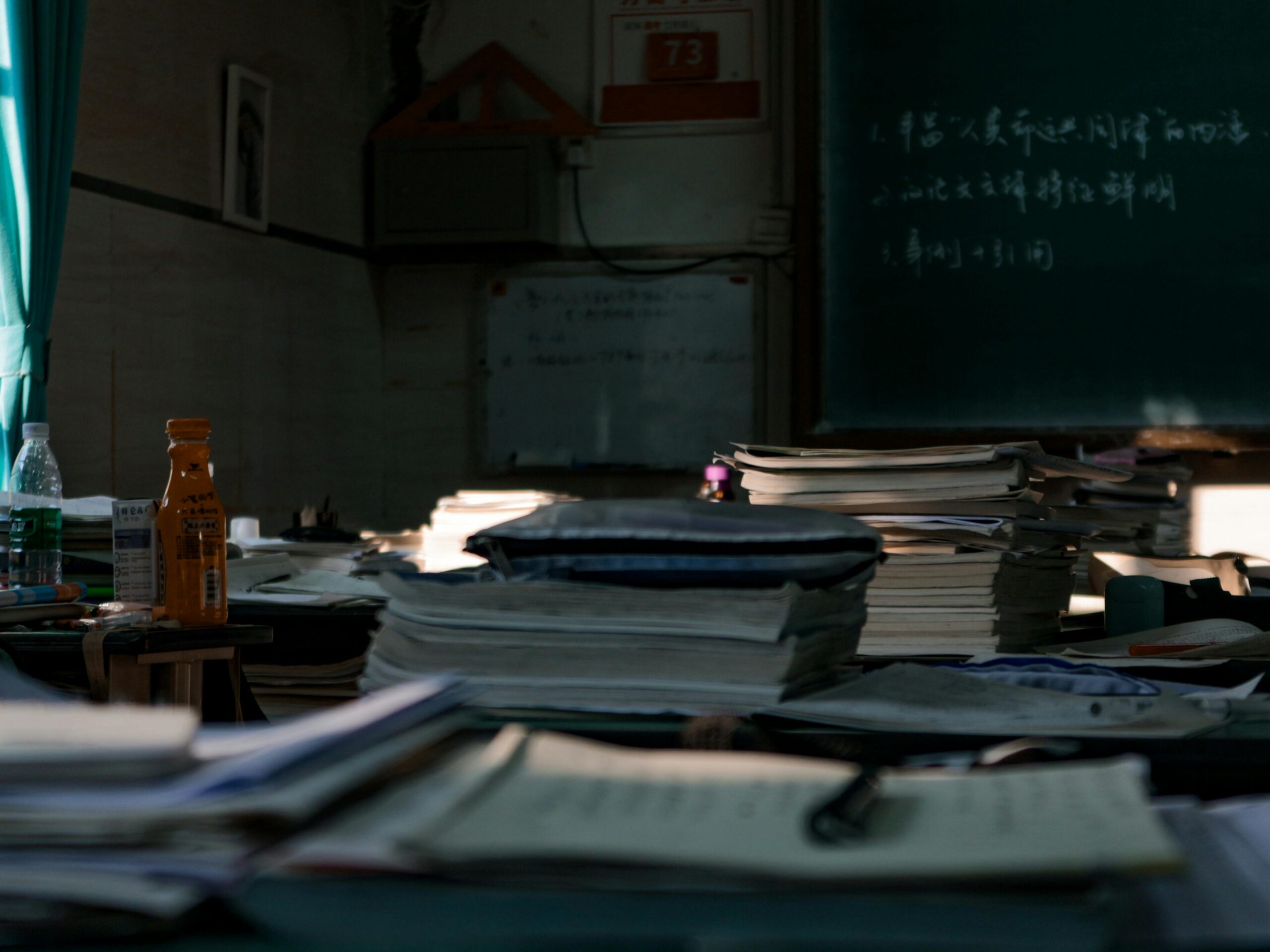 Messy tables with big heaps of papers and stationery in dark room with blackboard