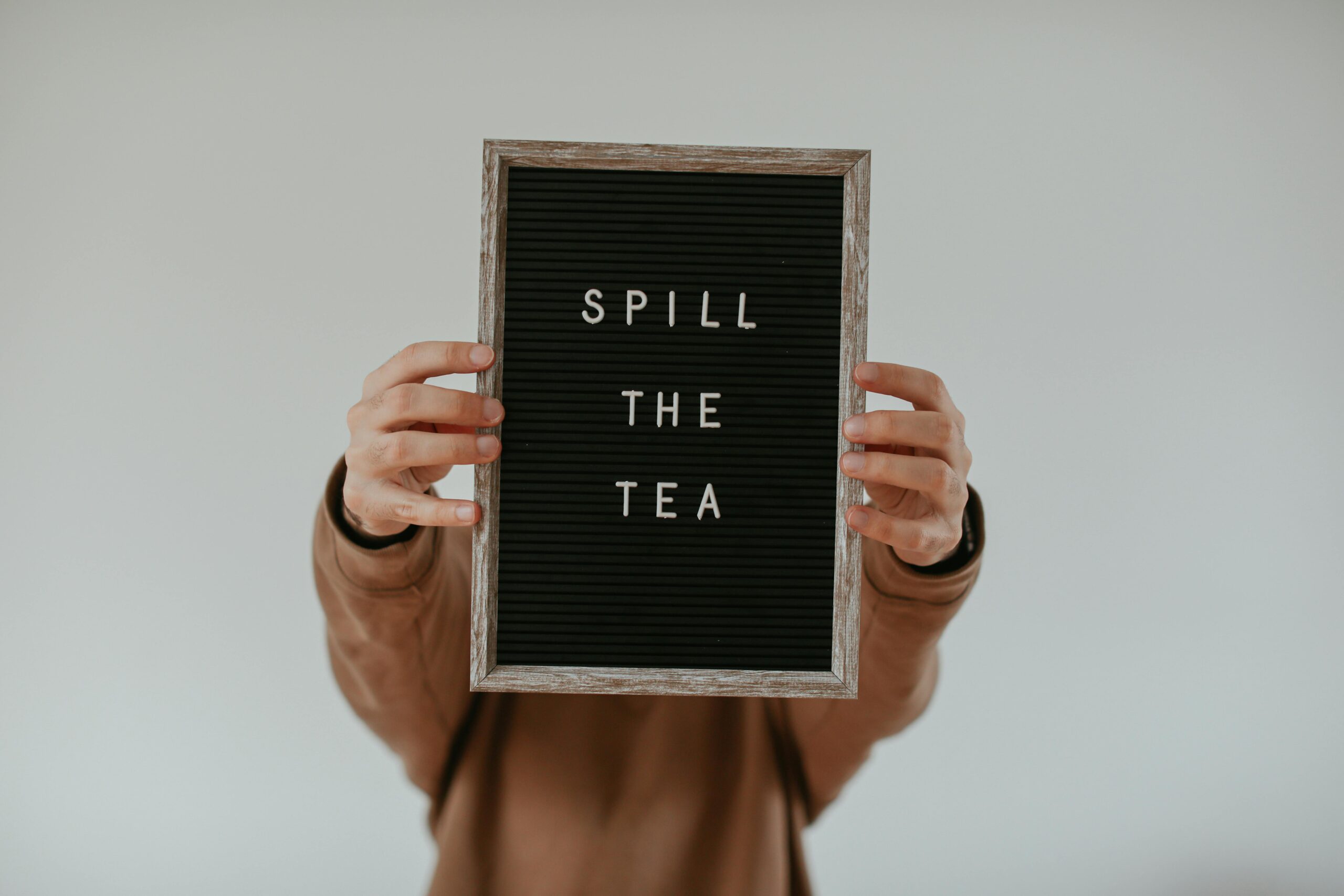 Person holding a letter board with the phrase 'Spill the Tea' against a white wall.