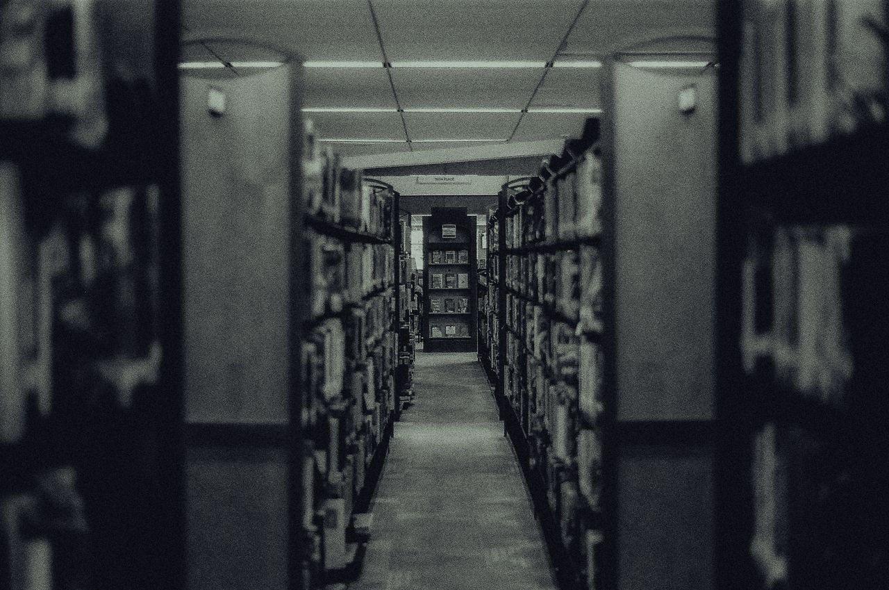 Atmospheric black and white photo capturing a library aisle with rows of shelves filled with books and a vanishing perspective.